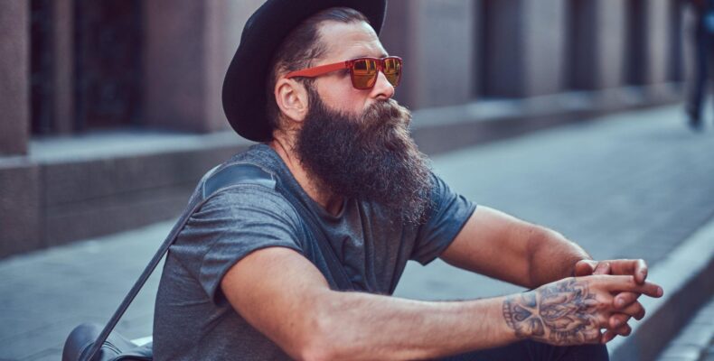 A bearded man in a hat and glasses sits on a sidewalk