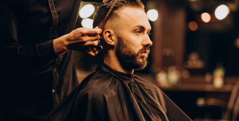 A barber trims the hair of a bearded man in a dimly lit salon.