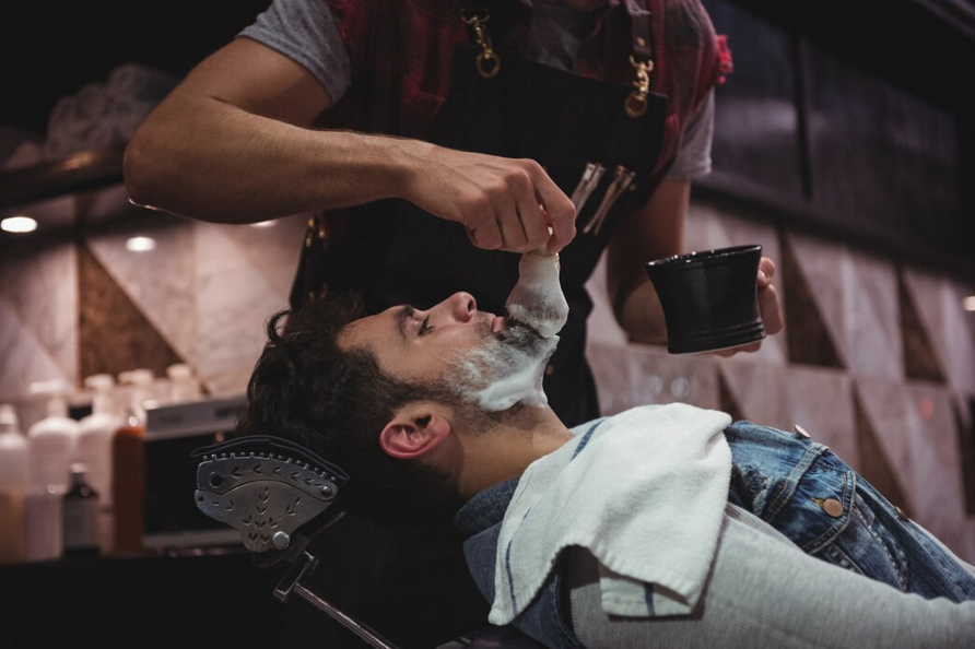 Man getting a traditional shave; barber applies foam with brush in a classic salon.